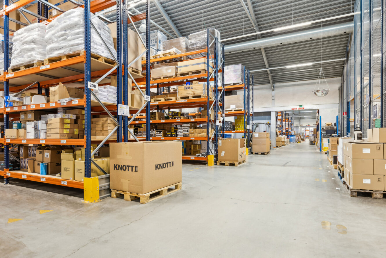 Lageret er indrettet med høje reoler fyldt med kasser og pakker, nogle på paller, organiseret langs brede, rene gange under et metalloft.