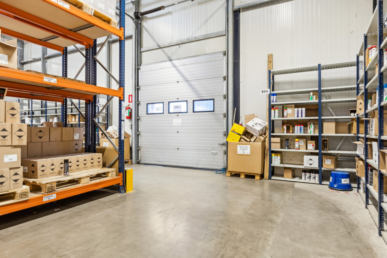 Industrielt lagerinteriør med hylder og paller fyldt med kasser og beholdere; en stor port ses i baggrunden.