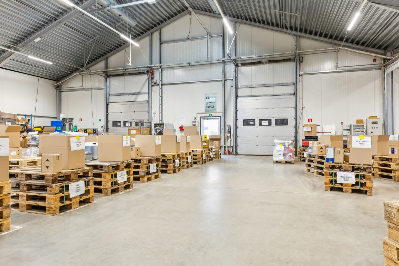 Lageret er indrettet med store papkasser stablet på træpaller i rækker på et betongulv under et metaltag.