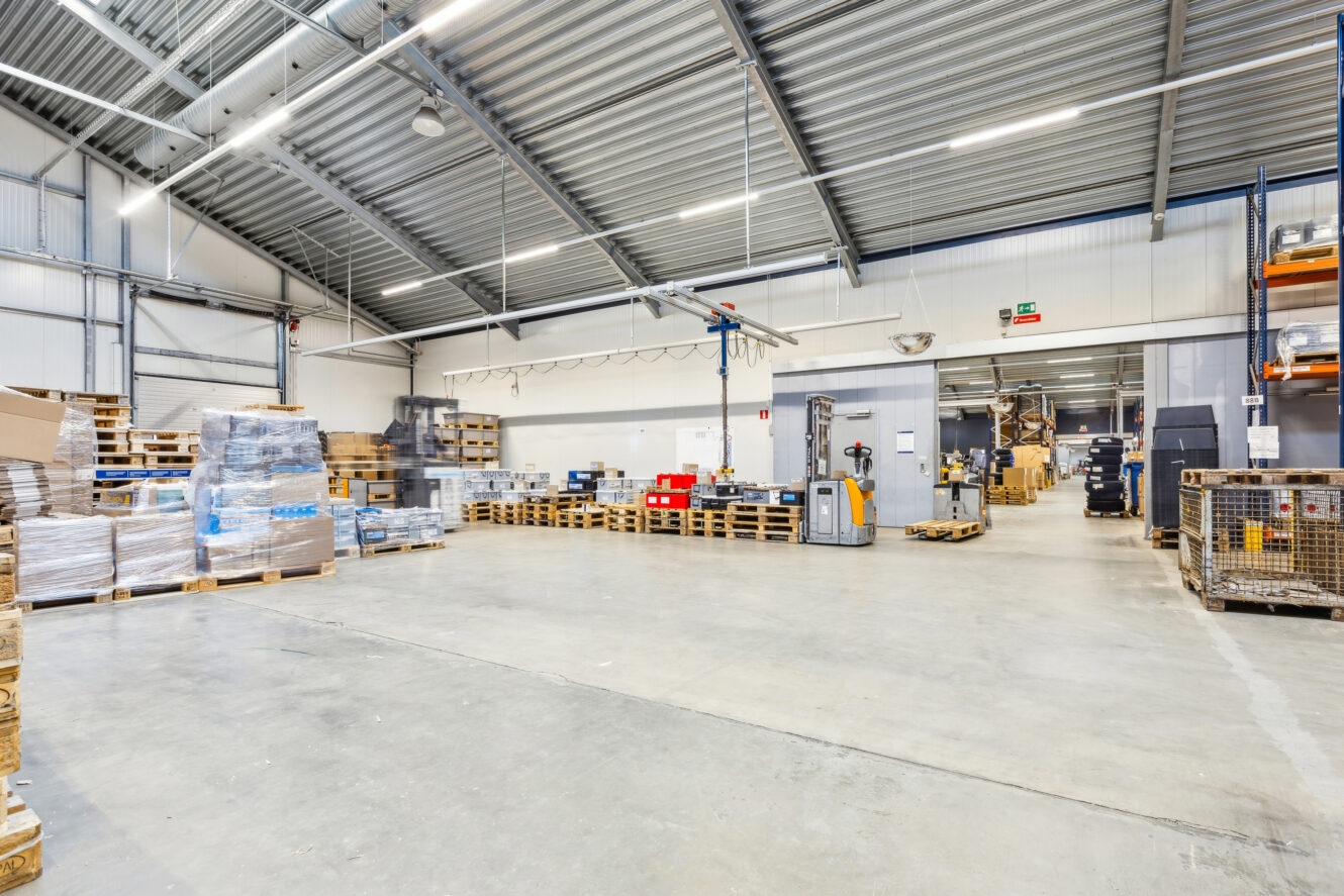 Rummeligt lagerinteriør med paller, kasser og udstyr arrangeret langs væggene under stærk belysning og et højt metalloft.