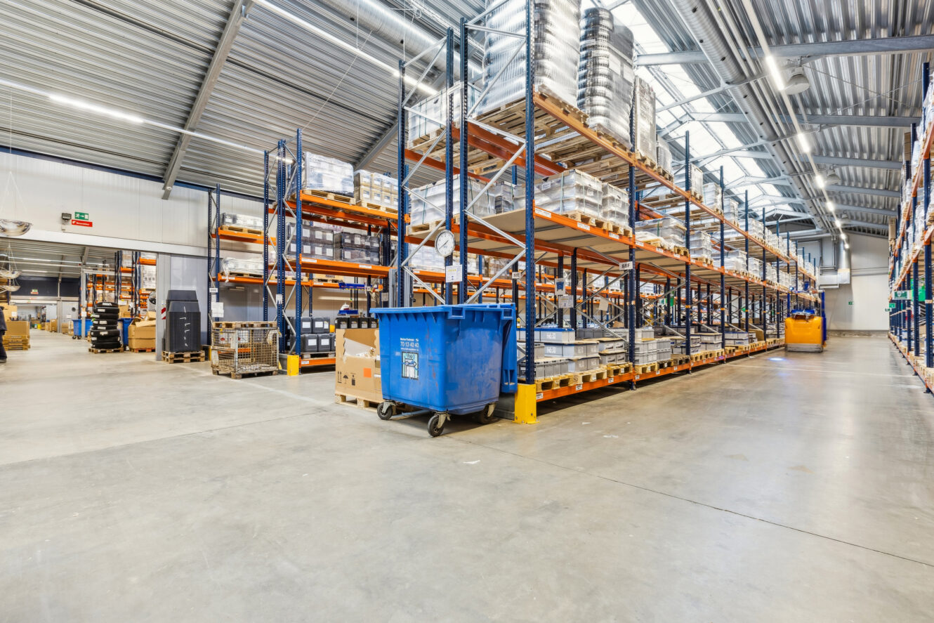 Rummelig lagerindretning med høje metalhylder fyldt med kasser og materialer, en blå genbrugsspand og kraftig ovenlys.