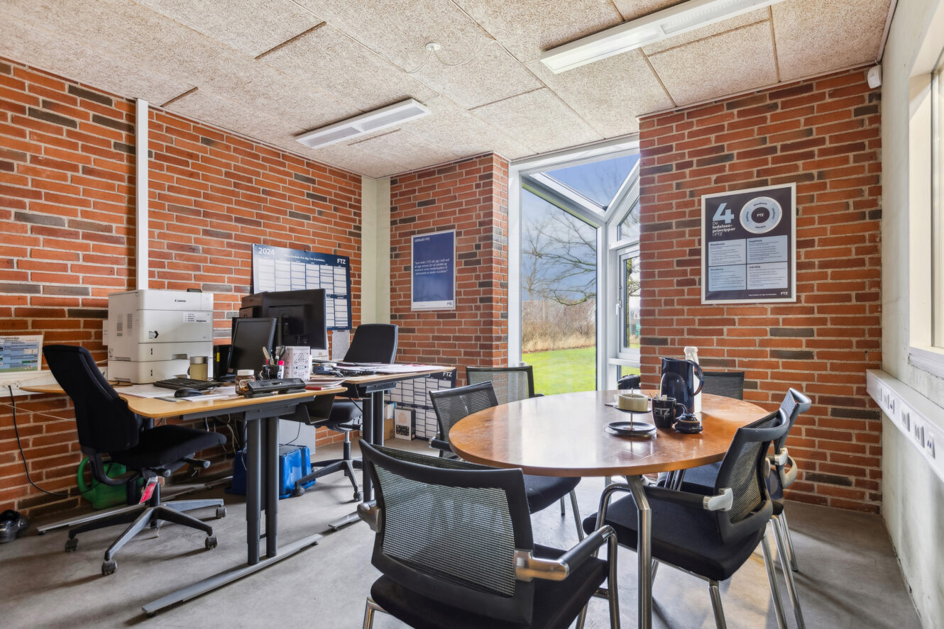 Kontor med murstensvægge, to skriveborde med computere og papirer, en printer, et rundt mødebord med stole og store vinduer, der lukker naturligt lys ind.