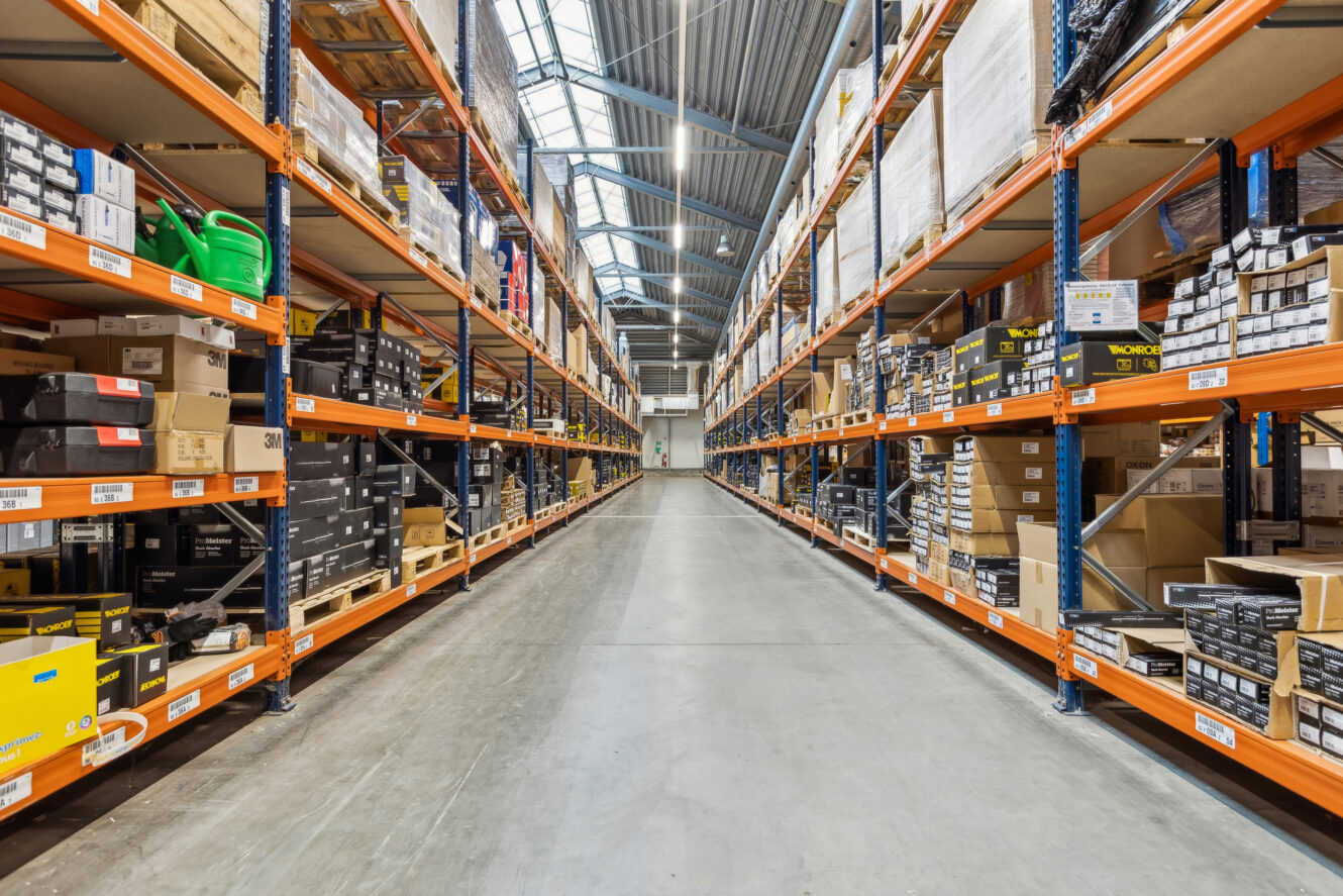 Bred lagergang med høje hylder på begge sider, fyldt med forskellige kasser og varer under kraftig ovenlys.