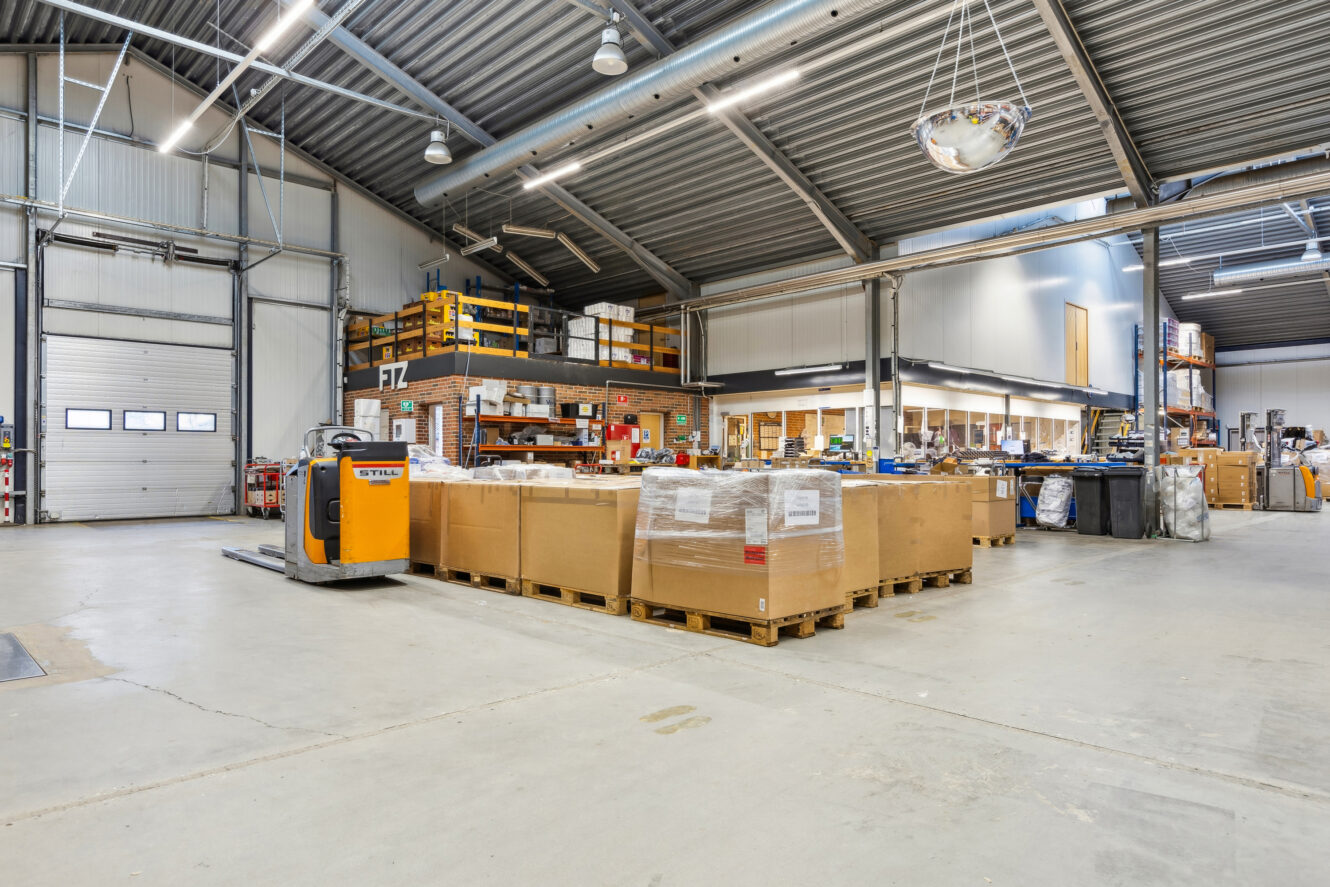 Et rummeligt lagerinteriør med paller med kasser, en palleløfter, høje hylder og en mezzanin med ekstra lager- og kontorplads.