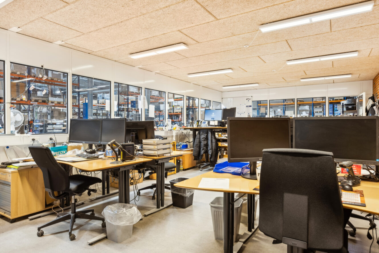 Moderne kontorlokaler med flere skriveborde, computerskærme og kontorstole; store vinduer afslører et industri- eller lagerområde i baggrunden.