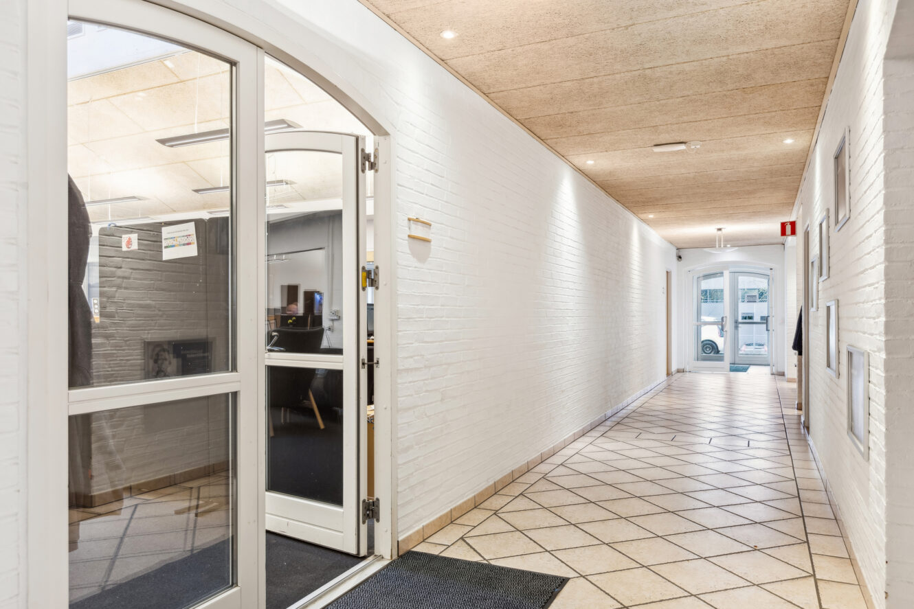 En ren gang med hvide murstensvægge, klinkegulv og en glasdør, der fører ind til et kontorlokale; en anden glasdør er synlig i den fjerneste ende.