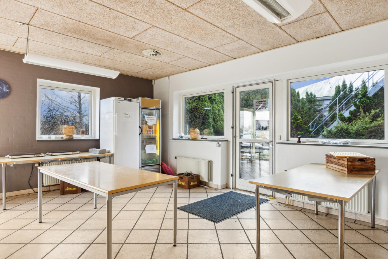 Et rent, lyst rum med klinkegulv, to rektangulære borde, et køleskab og store vinduer med udsigt til de grønne områder udenfor.