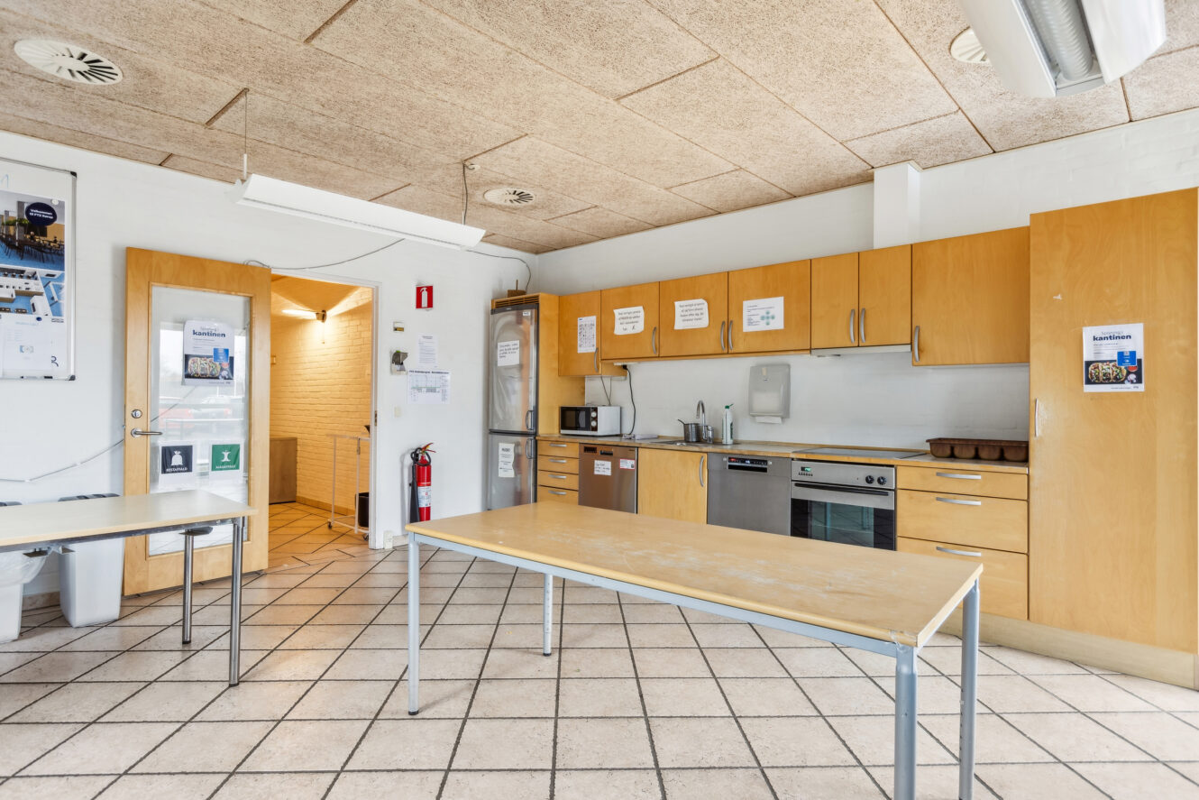Et rent fælleskøkken med træskabe, klinkegulv, to borde og forskellige apparater, herunder ovn, mikroovn og vask. Der er sikkerheds- og instruktionsskilte på vægge og skabe.