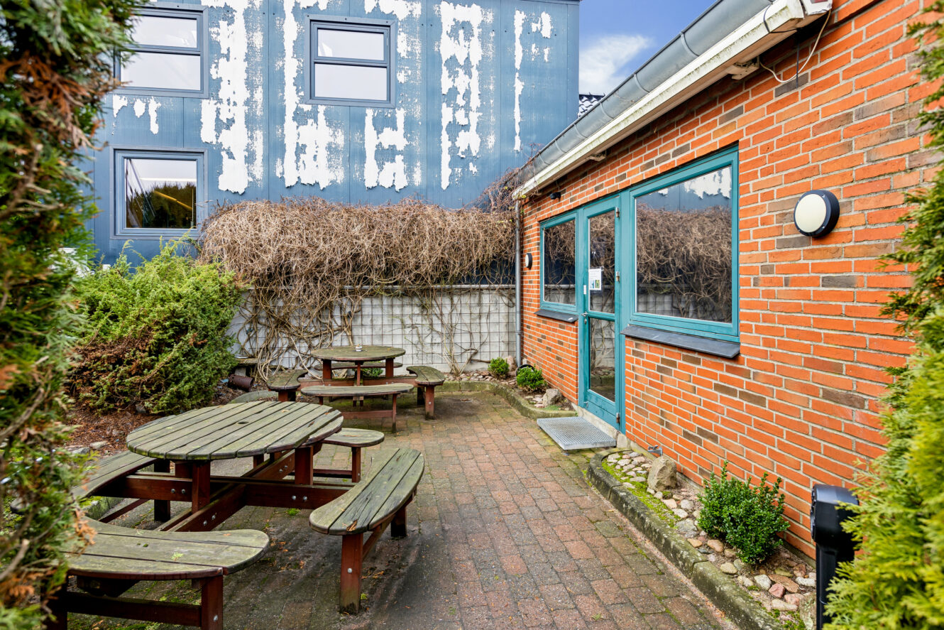 Lille muret gårdhave med picnicborde af træ, omgivet af buske, ved siden af en murstensbygning. En forvitret blå bygning med afskallet maling ses i baggrunden.