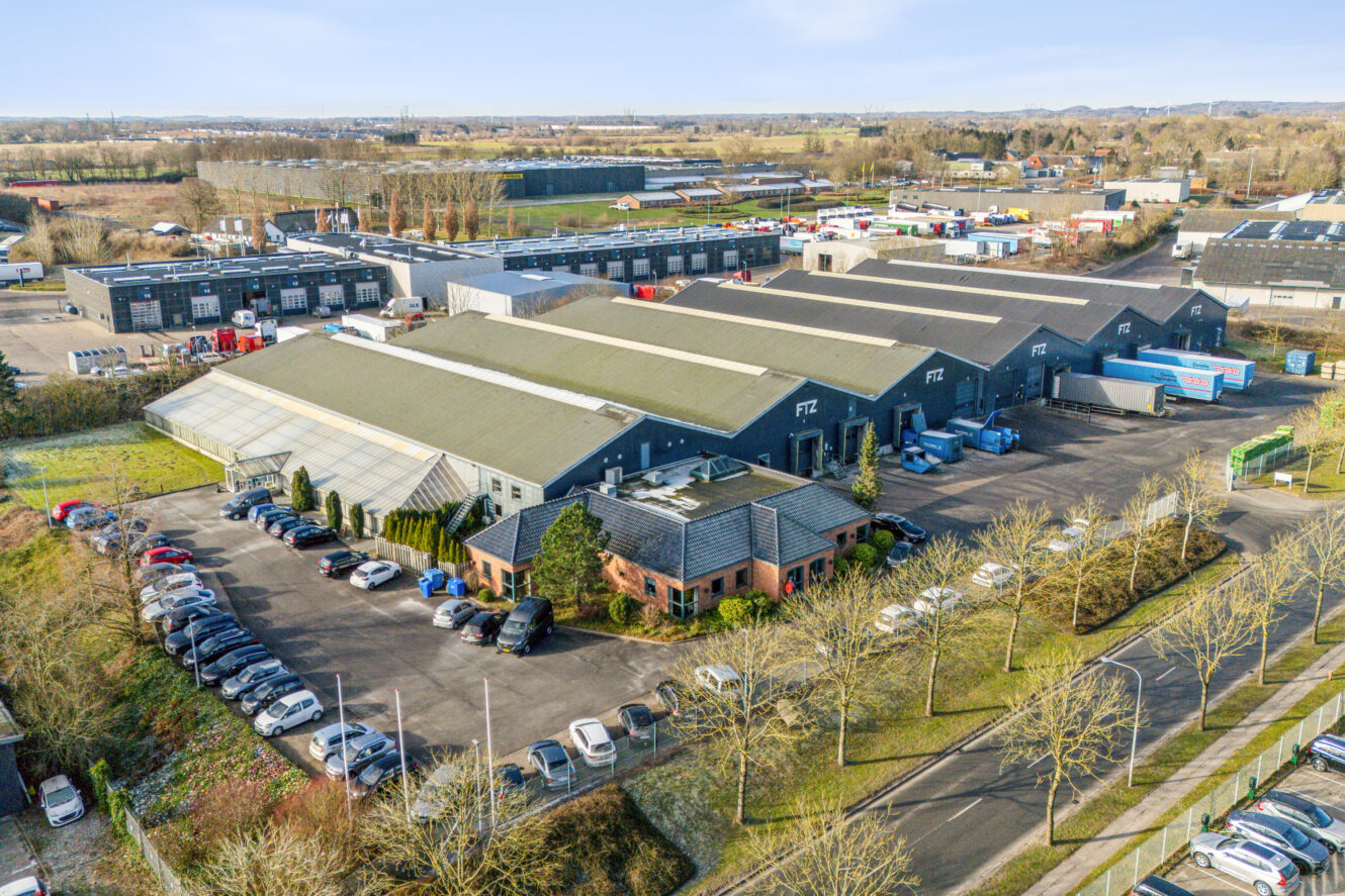 Luftfoto af et industrikompleks med store lagerbygninger, en bilfyldt parkeringsplads og træer og veje omkring området.