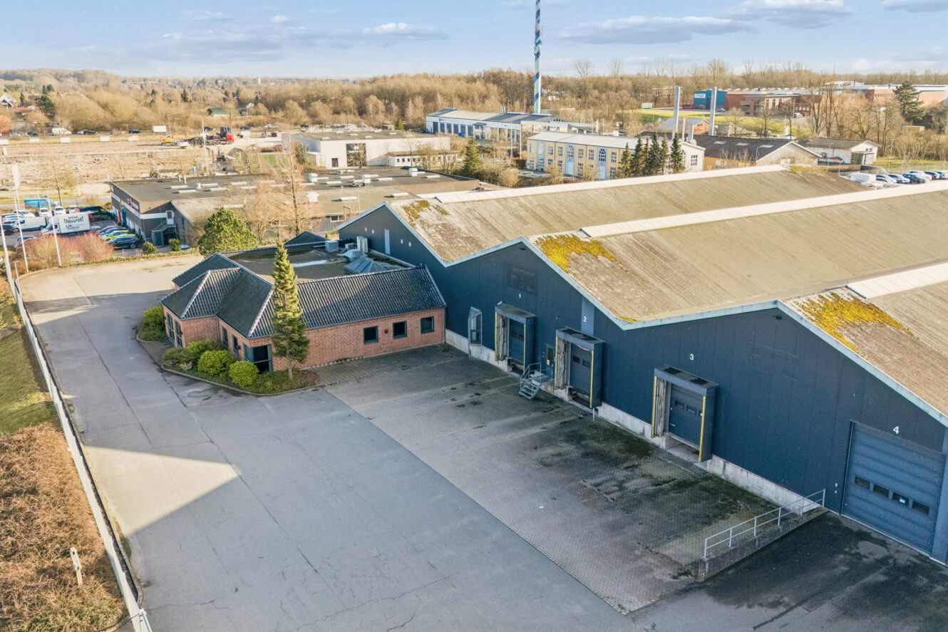 Et stort industrilager med læsseramper, en kontorbygning i mursten og en tom grund omgivet af andre bygninger og træer.