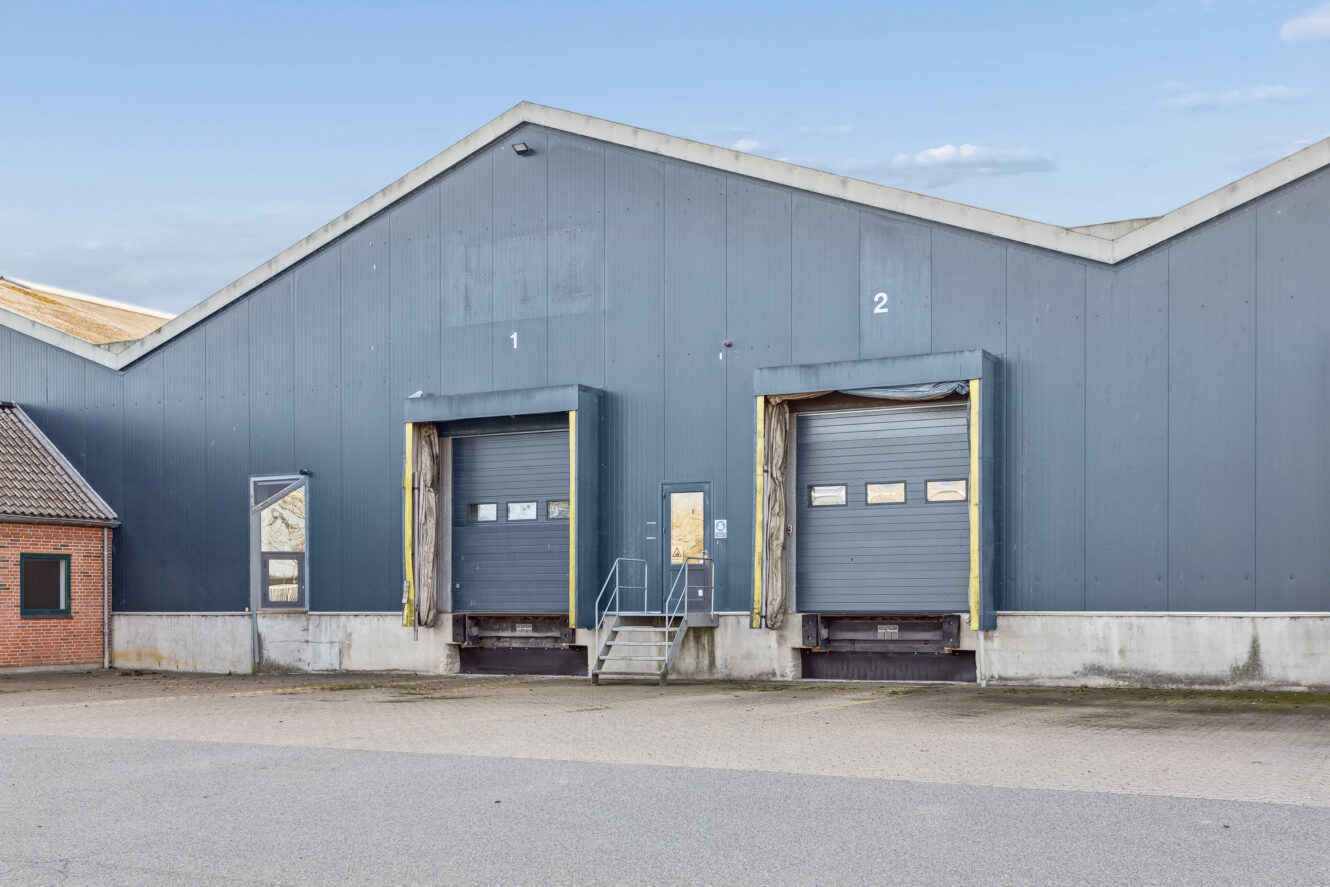 Grå industribygning med to læsseramper mærket 1 og 2, lukkede rulleporte og metaltrapper på forsiden.