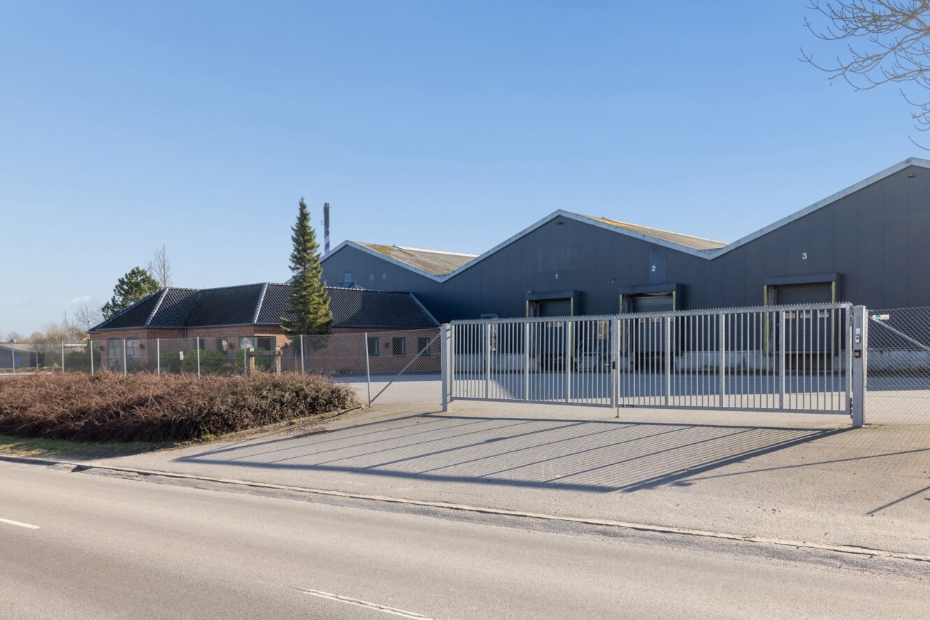 Lukket indgang til et industrielt lagerkompleks med læsseramper og et muret kontor, set fra den anden side af en tom vej på en klar dag.