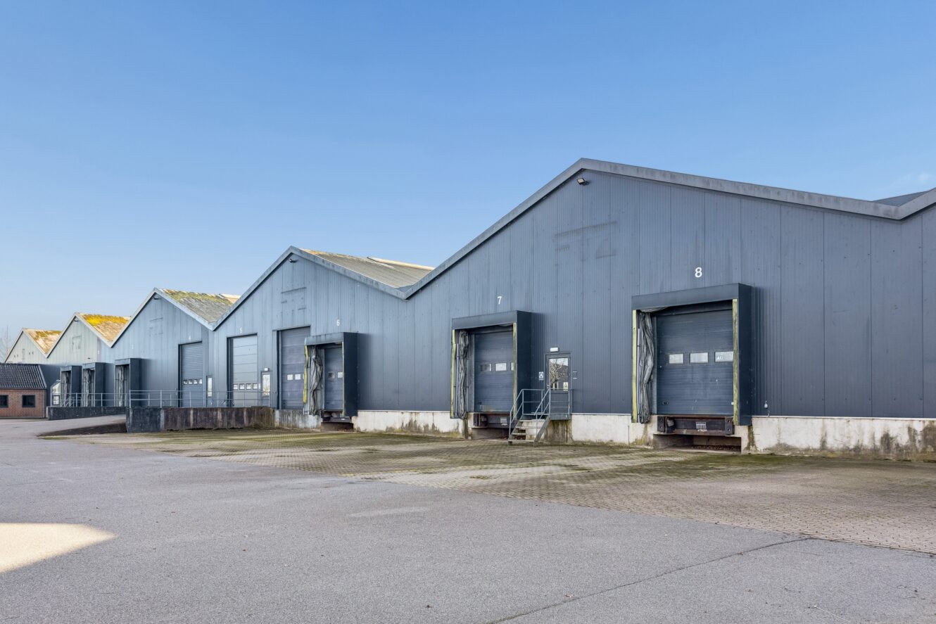 En række grå industrihaller med læsseramper og lukkede porte under en klar blå himmel.