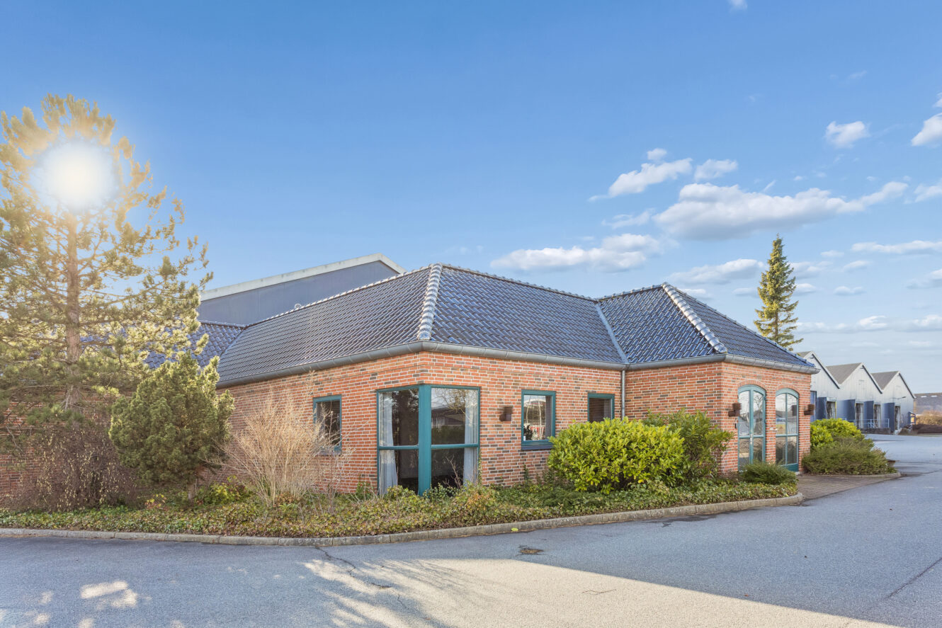 Rød murstensbygning i én etage med mørke tagsten, omgivet af buske og træer, under en klar himmel med spredte skyer.