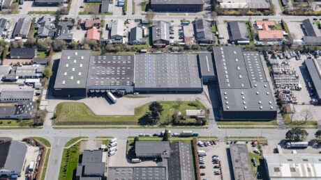 Luftfoto af et stort industrielt bygningskompleks omgivet af veje, parkerede køretøjer og nærliggende beboelses- og erhvervsbygninger.