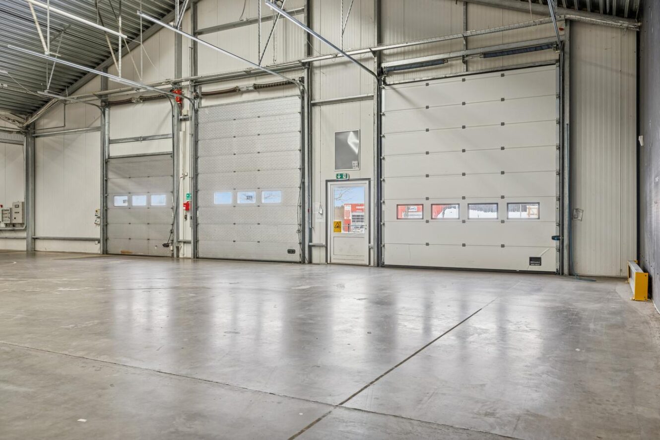 Tomt industrilager med betongulv, højt til loftet og tre store rullende garageporte langs den ene væg.
