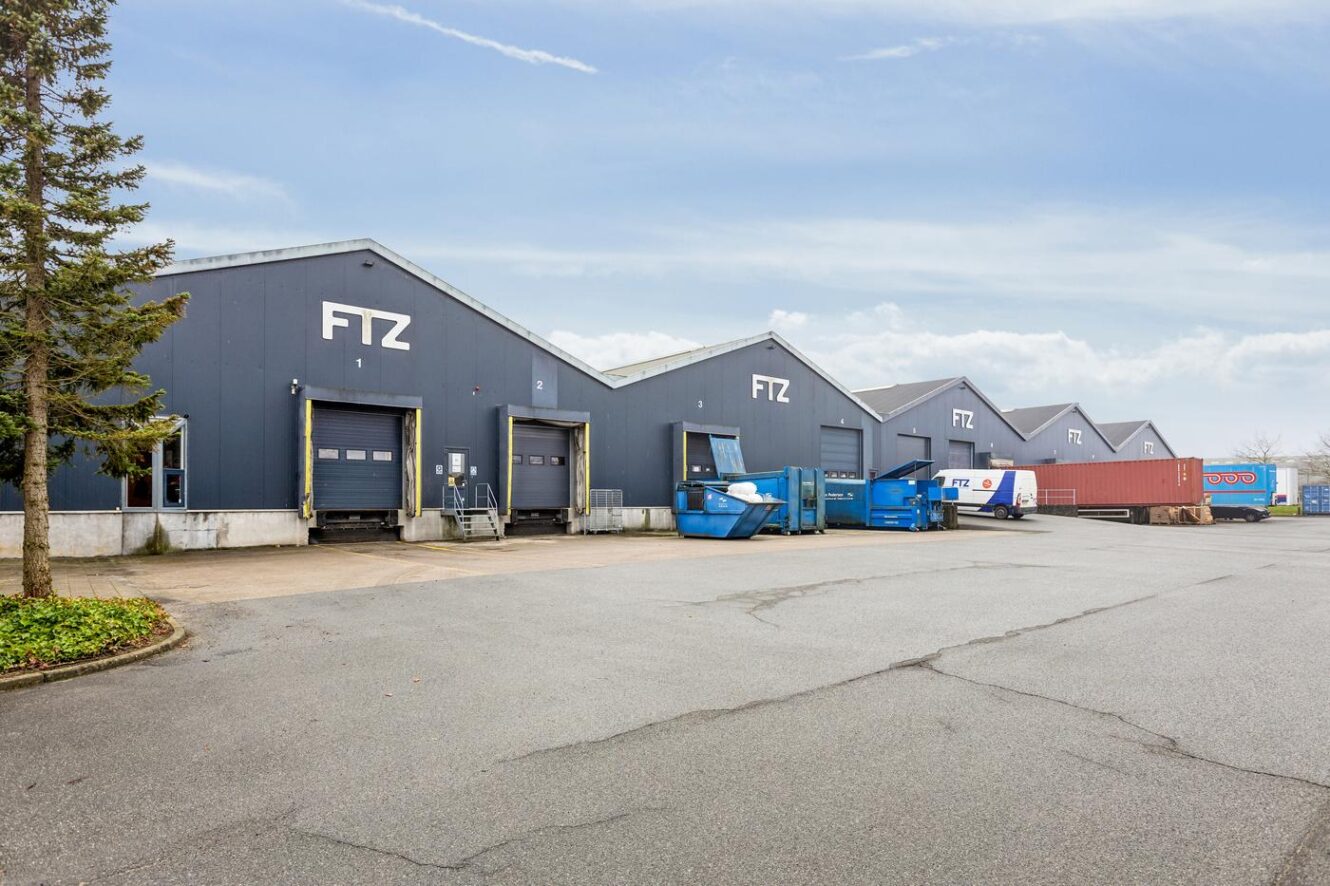 Udsigt til en lagerbygning mærket FTZ med flere læsseramper, containere og lastbiler parkeret udenfor under en overskyet himmel.