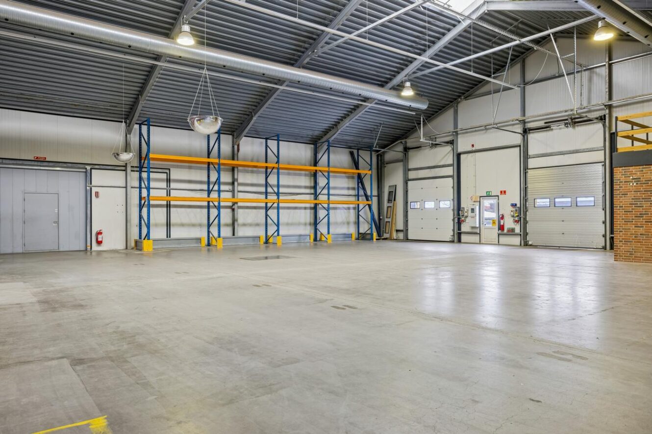 Interiør af et tomt industrilager med metalreoler, betongulv, metalloft, store garageporte og fluorescerende belysning.