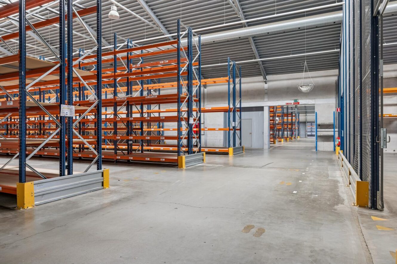 Tomt industrilager med metalreoler, betongulv og højt til loftet med synlige bjælker og lys.