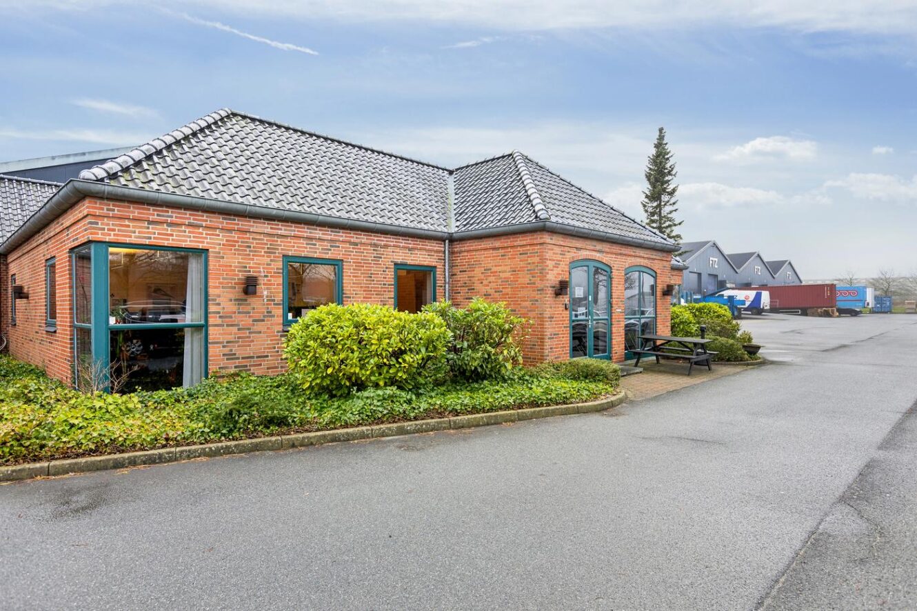 Murstensbygning i én etage med tegltag, store vinduer, omgivende buske og asfalteret parkeringsplads under en overskyet himmel.