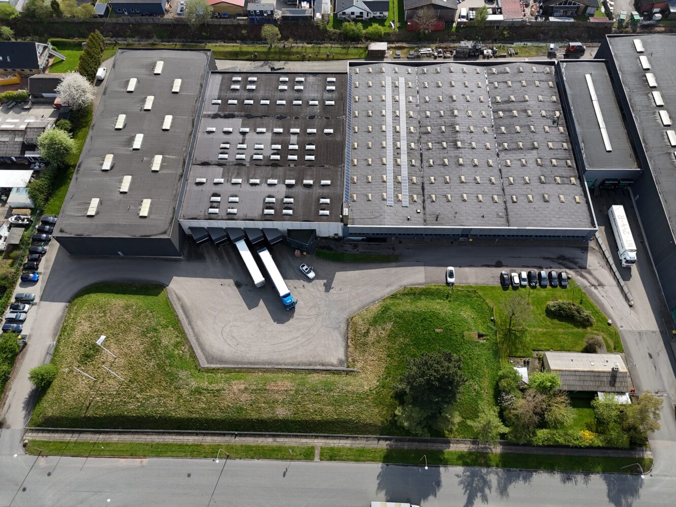 Luftfoto af en stor lagerbygning med flere læsseramper, parkerede lastbiler, biler og grønne områder omkring bygningen.