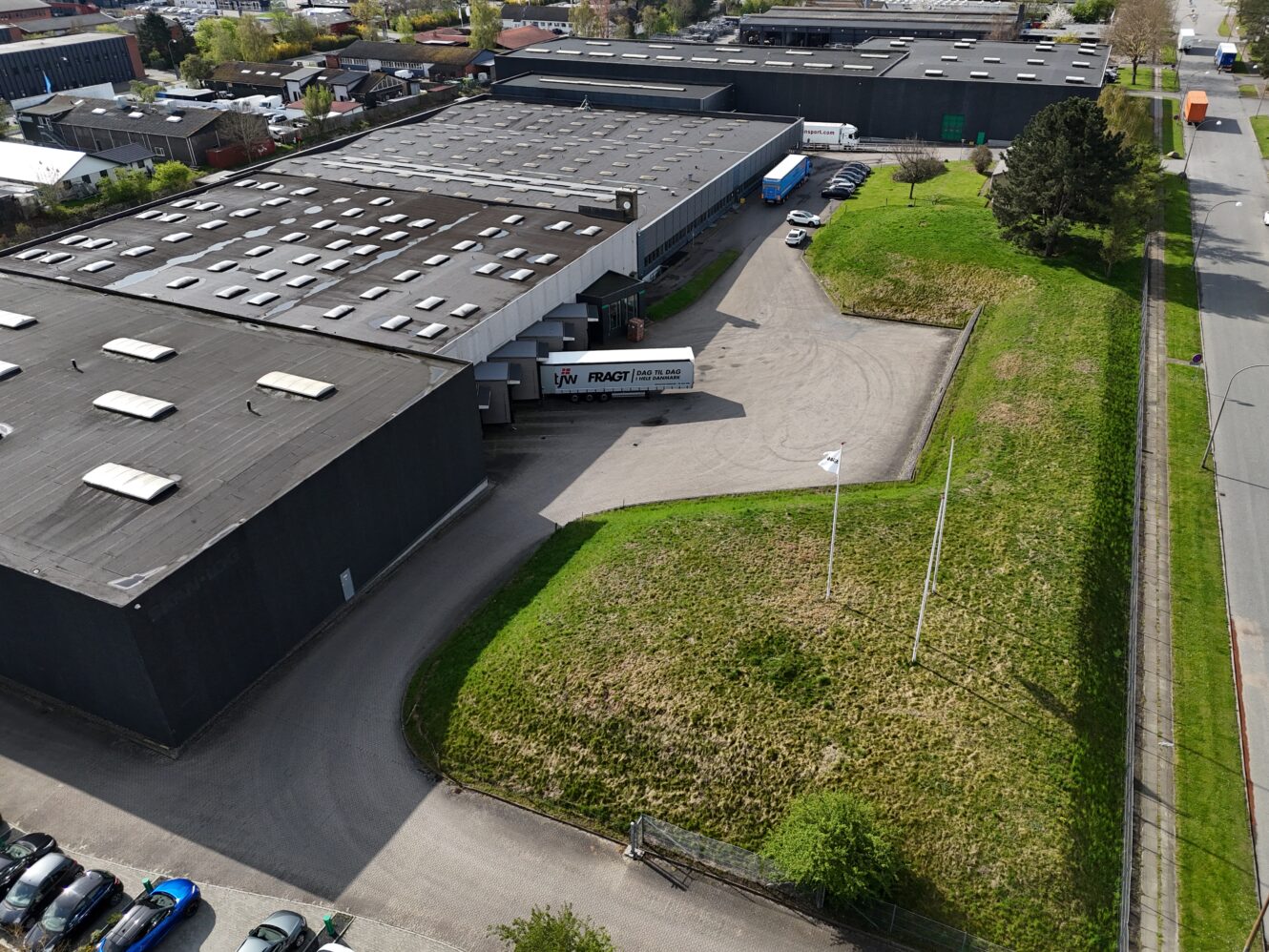 Luftfoto af et stort industrilager med en lastbil parkeret ved en læsserampe, der støder op til et græsareal og en parkeringsplads med flere biler.