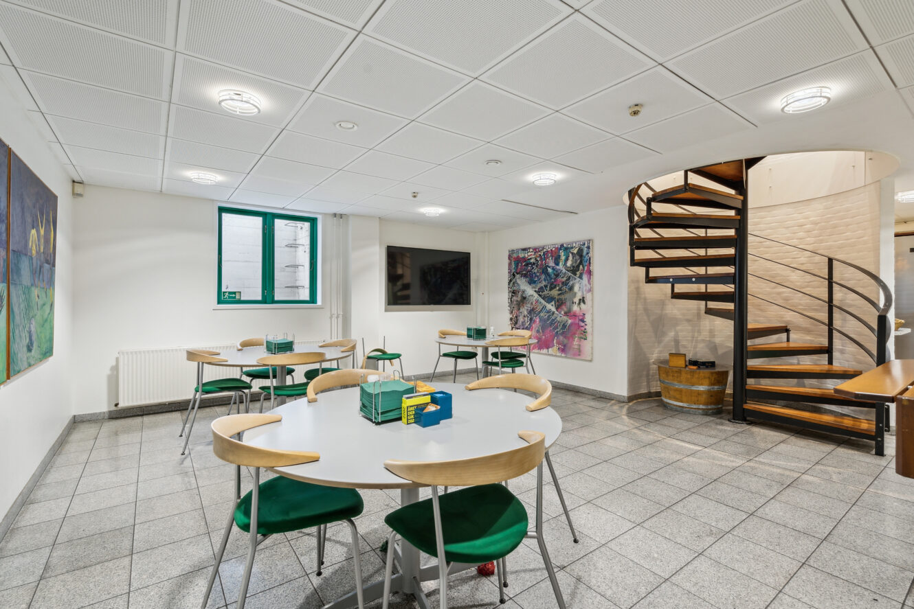 Et moderne rum med runde borde og grønne stole, abstrakte kunstværker på væggene, et fladskærms-tv og en vindeltrappe, der fører opad.