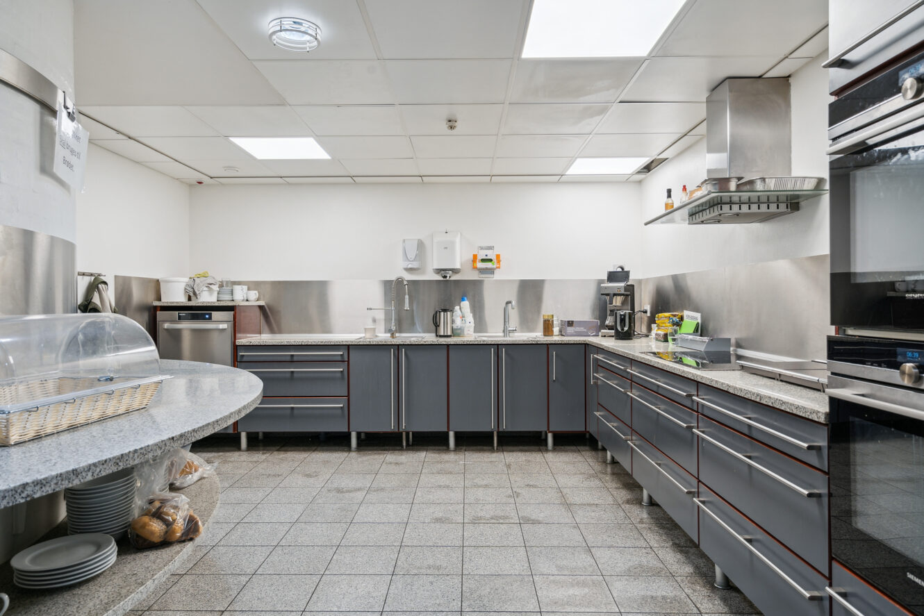 Moderne storkøkken med hvidevarer i rustfrit stål, grå skabe, bordplader og klinkegulv; tallerkener og brød står på et rundt sidebord.