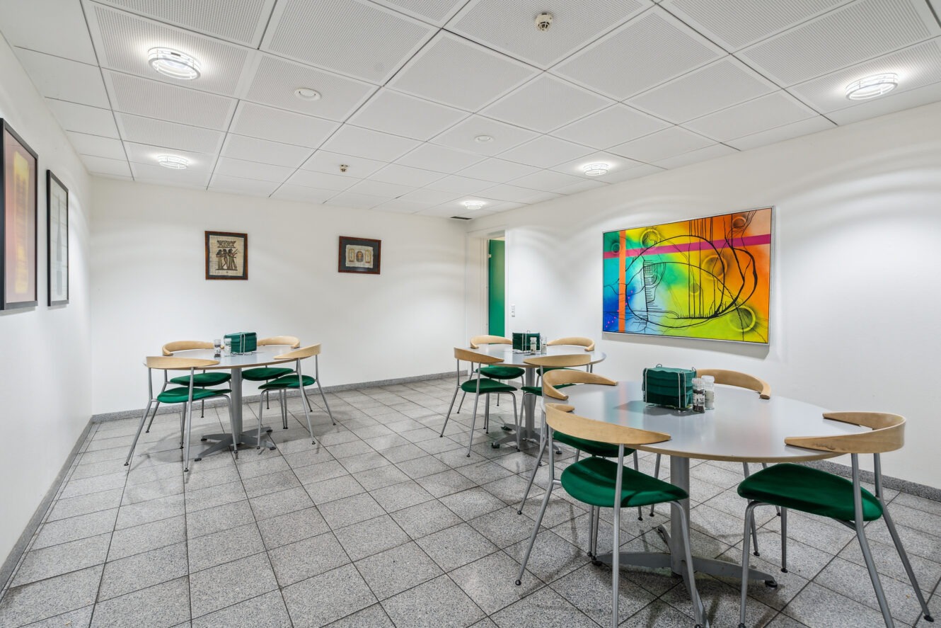 Et lille cafeteria med tre runde borde, grønne stole, klinkegulv, vægkunst og kraftig ovenlys.