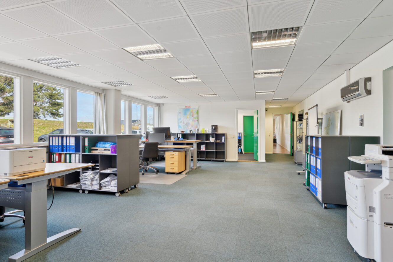 Moderne åbent kontorlandskab med skriveborde, stole, hylder, arkivskabe og vinduer langs den ene væg, oplyst af loftslamper.