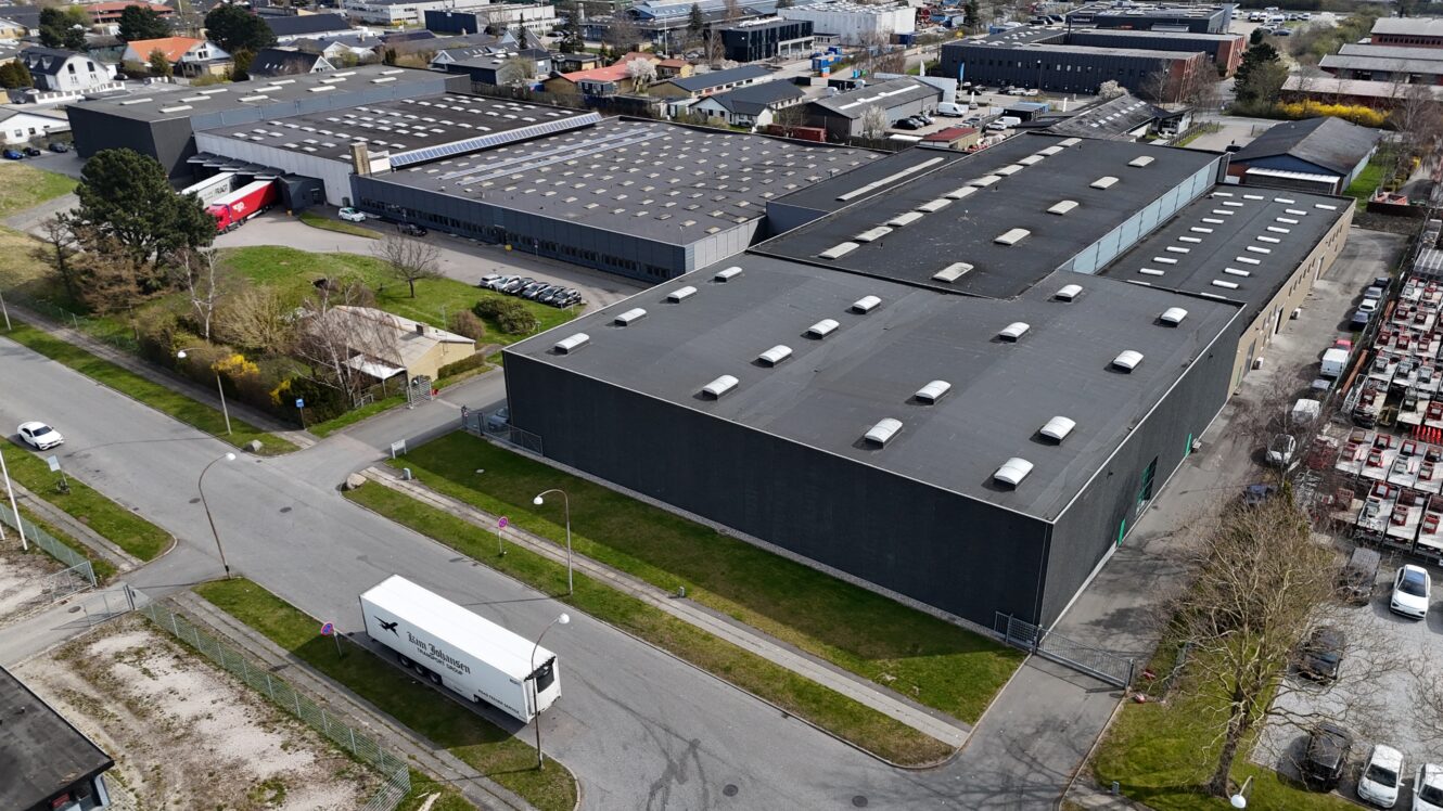 Luftfoto af store industribygninger med flade tage, omgivet af veje; en hvid lastbil er parkeret i nærheden, og andre kommercielle strukturer er synlige i baggrunden.