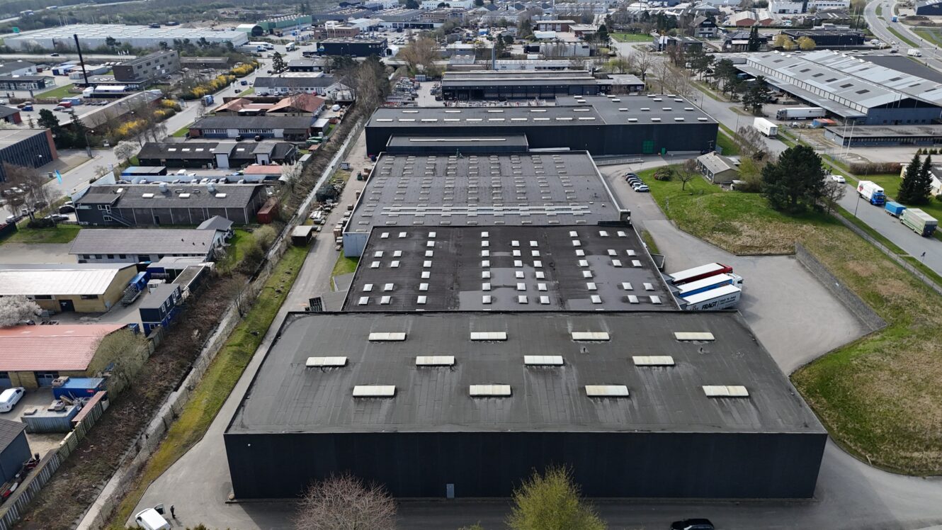 Luftfoto af flere store industrielle lagerbygninger med lastbiler parkeret i nærheden, omgivet af veje og andre erhvervsbygninger.