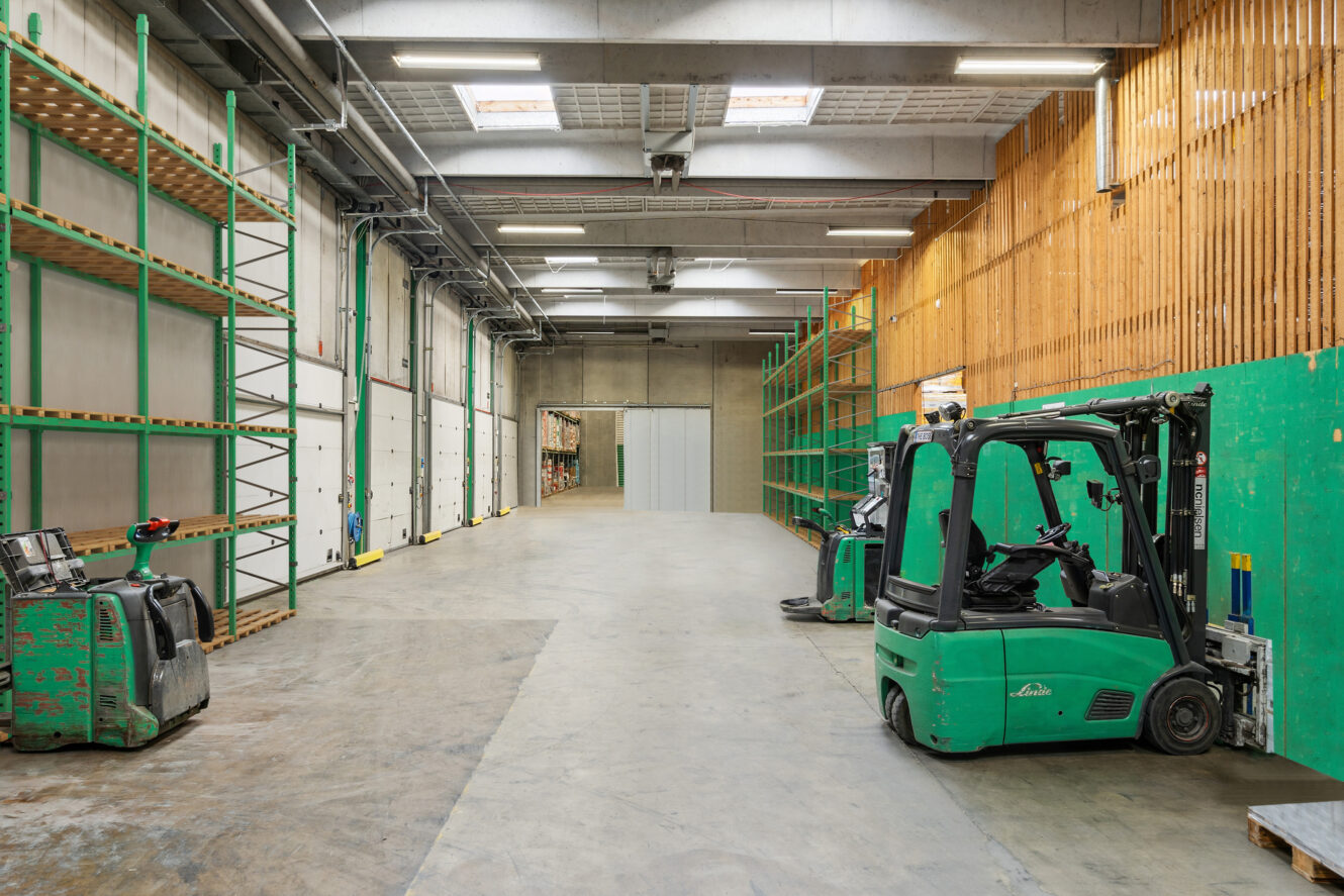 En stor, tom lagerhal med grønne metalreoler, palleløftere og gaffeltrucks langs siderne under fluorescerende belysning.