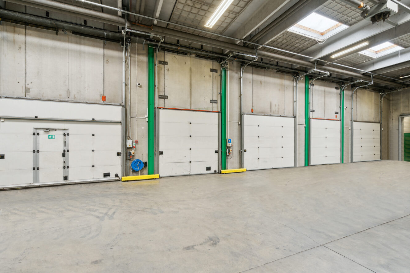 Stort, tomt industrilager med flere lukkede, hvide porte til læsseramper, betongulv, højt til loftet og ovenlys.