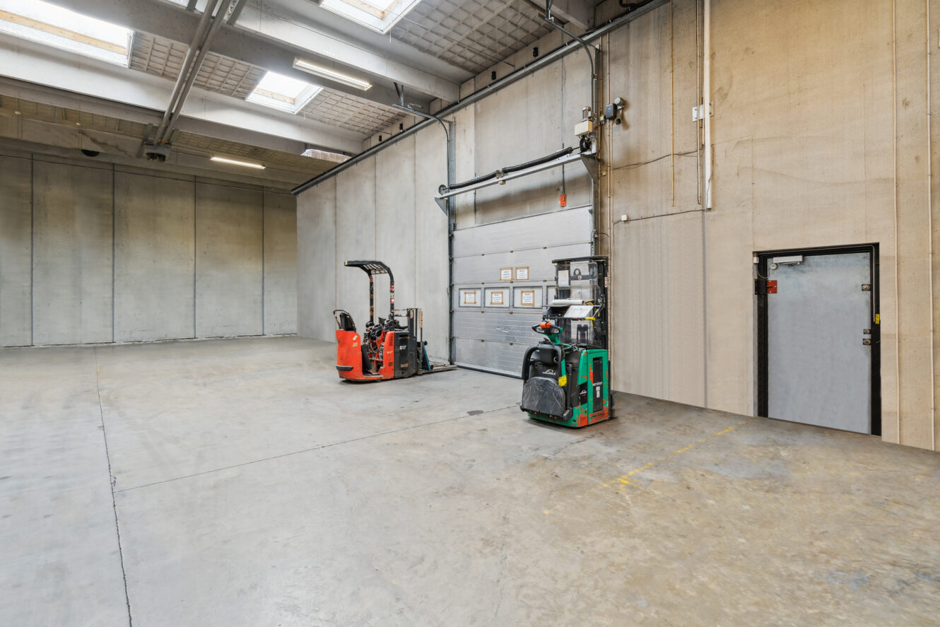 Rummeligt industrielt lagerinteriør med to palletrucks parkeret nær en lukket garageport og en sidedør på højre væg.