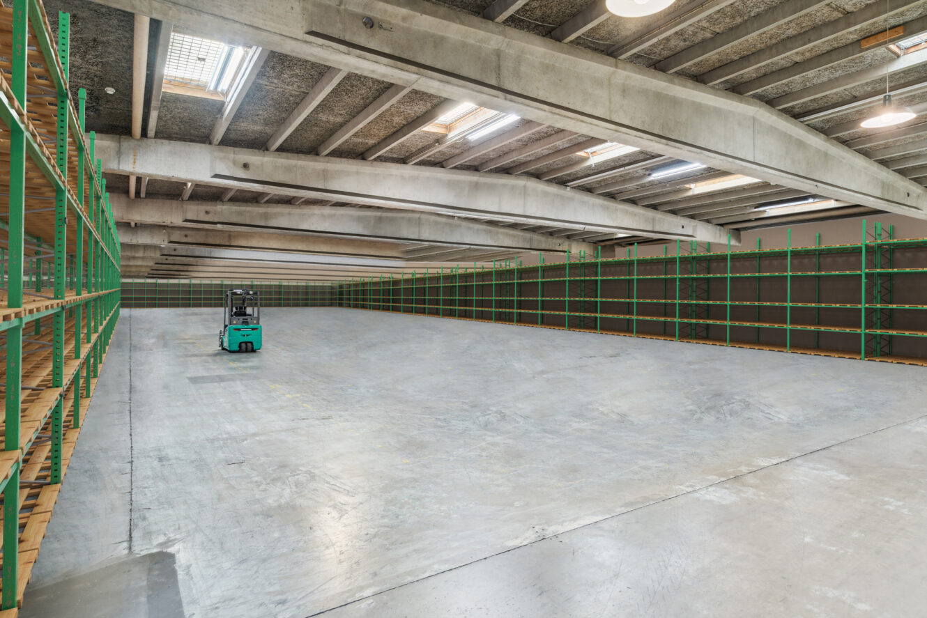 Stor tom lagerbygning med grønne metalreoler langs væggene og en enkelt blågrøn gaffeltruck parkeret på betongulvet.