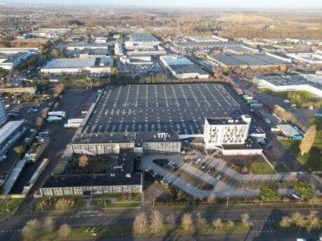 Luftfoto af et stort industrikompleks med parkeringsplads, omkringliggende veje og flere bygninger i et forstadsområde.
