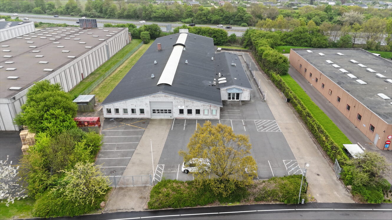 Luftfoto af en industribygning med mørkt tag, tilstødende parkeringsplads og omkringliggende grønne områder, beliggende mellem to andre store lagerbygninger.