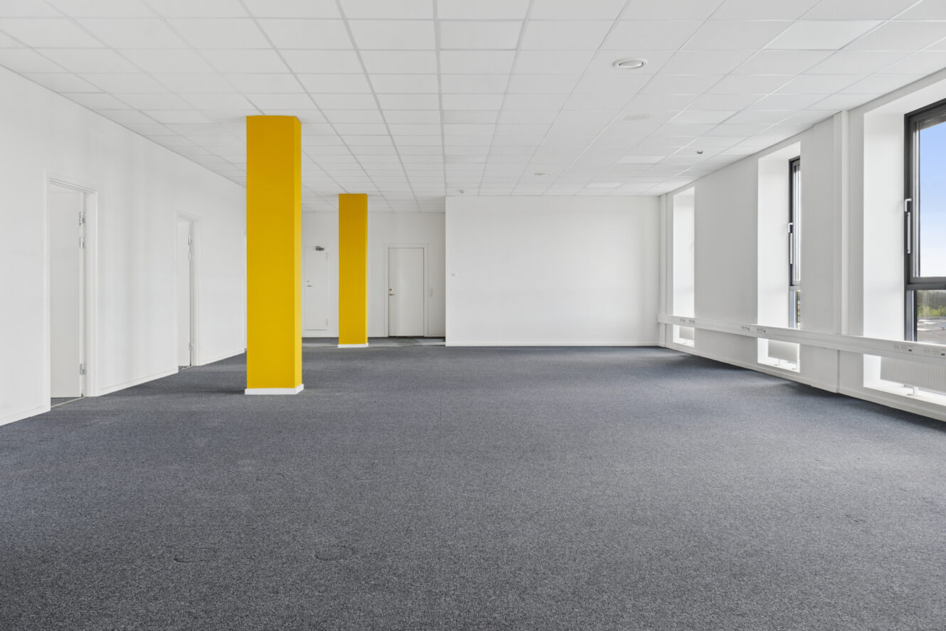Tomt kontorlokale med gråt gulvtæppe, hvide vægge, store vinduer og to lysegule søjler.