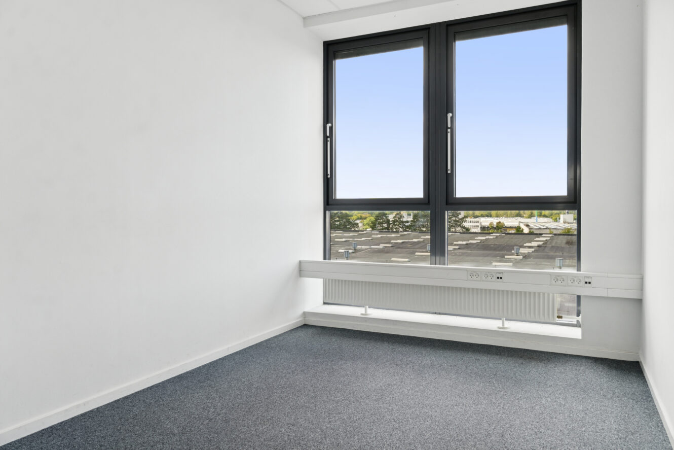 Lille tomt kontorlokale med hvide vægge, blåt gulvtæppe, stort vindue, radiator og stikkontakter under vinduet med udsigt til et fladt tag og træer i det fjerne.