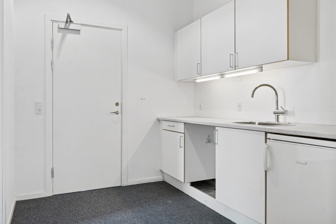 Et lille hvidt tekøkken med over- og underskabe, en vask, en bordplade og en lukket dør til venstre. En skabslåge under vasken mangler.
