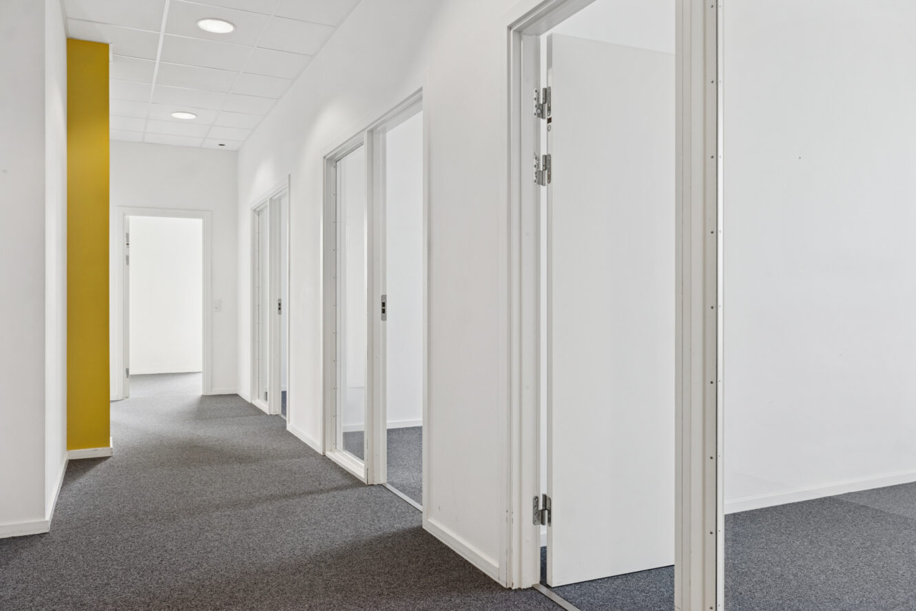 En moderne kontorgang med gråt gulvtæppe, hvide vægge, flere døråbninger, der fører til tomme rum, og en gul accentvæg til venstre.