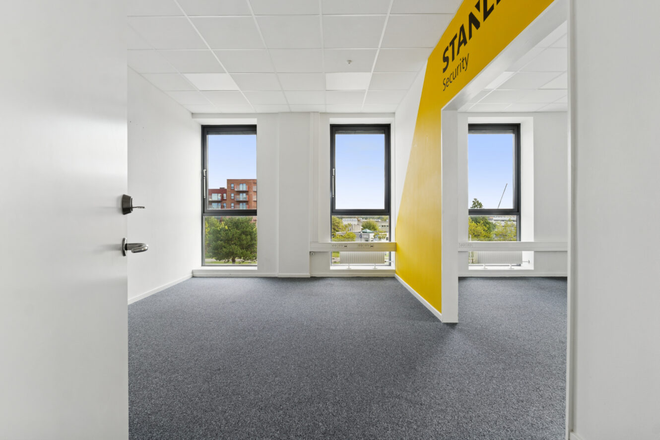 Et moderne, umøbleret kontorlokale med gråt gulvtæppe, hvide vægge, store vinduer og en gul skillevæg med ordet STANLEY.