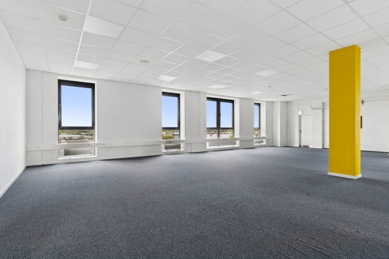 Tomt kontorlokale med gråt gulvtæppe, hvide vægge, tre store vinduer, en gul søjle og et hvidt gitterloft.