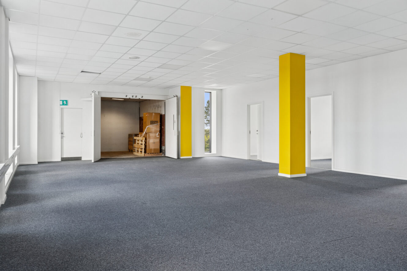 Tomt kontorlokale med gråt gulvtæppe, hvide vægge, gule søjler, flere døre og en stak kasser i nærheden af et åbent læsseområde.