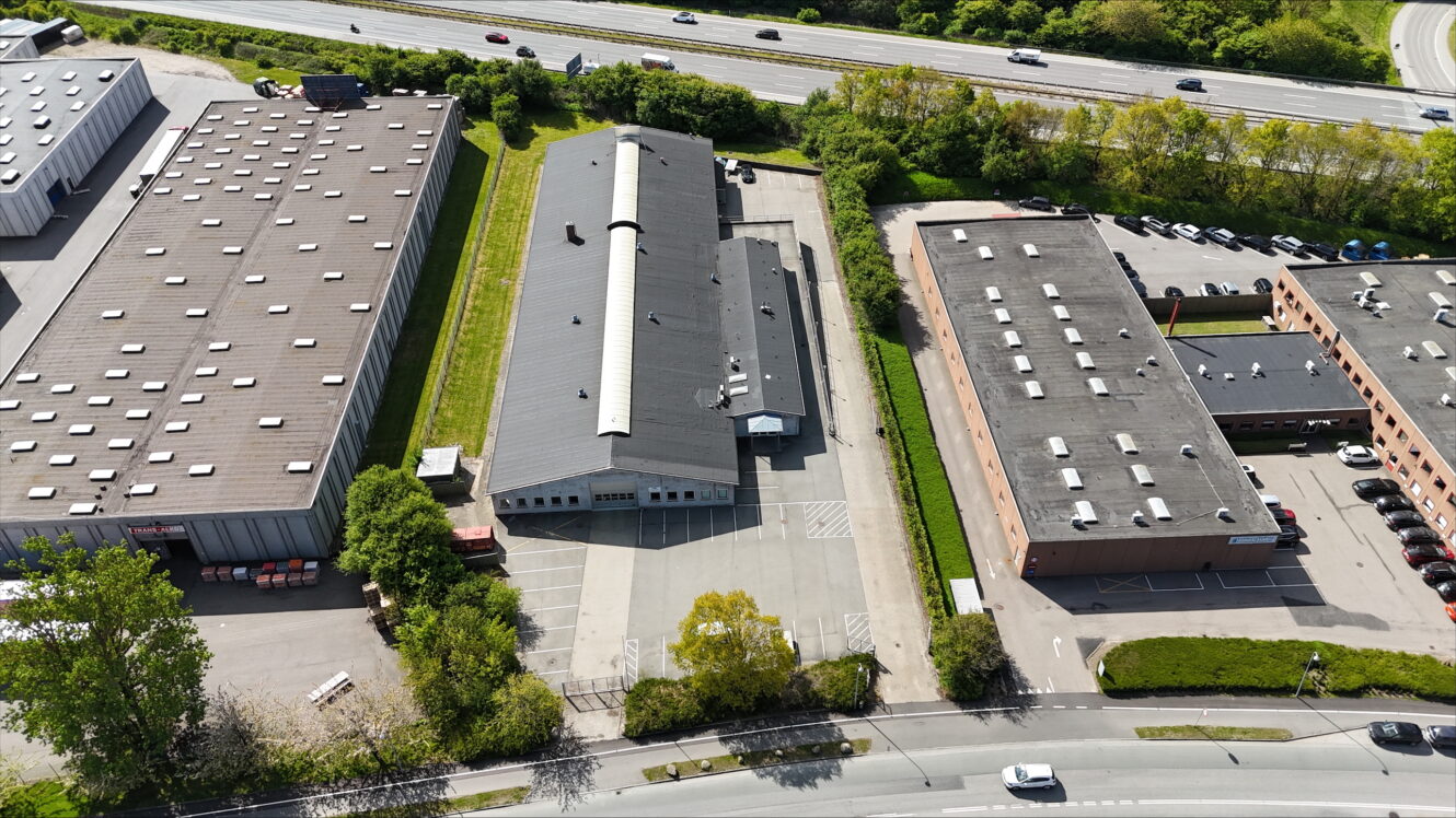 Luftfoto af tre industribygninger med parkeringspladser, omgivet af veje, grønne områder og køretøjer, i nærheden af en motorvej.