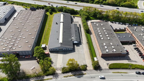 Luftfoto af tre industribygninger med parkeringspladser, omgivet af veje, grønne områder og køretøjer, i nærheden af en motorvej.