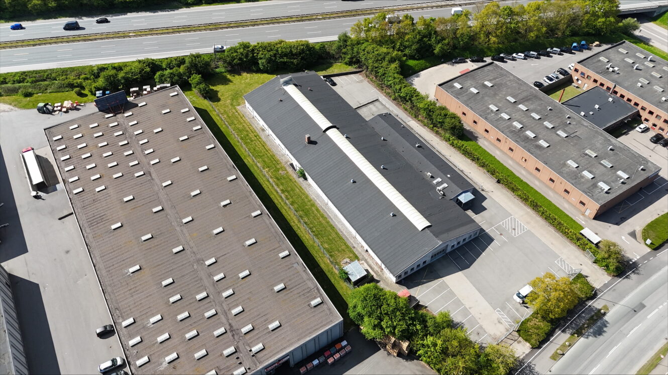 Luftfoto af industribygninger med flade tage, en parkeringsplads, grønne områder og en motorvej med kørende køretøjer i baggrunden.