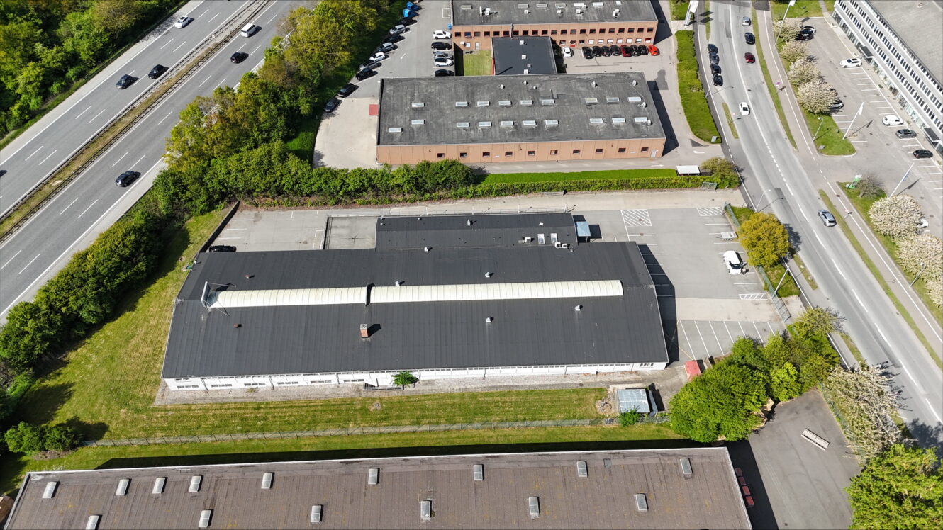 Luftfoto af industribygninger med parkeringspladser, omgivet af veje, grønne områder og nærliggende motorvejstrafik.
